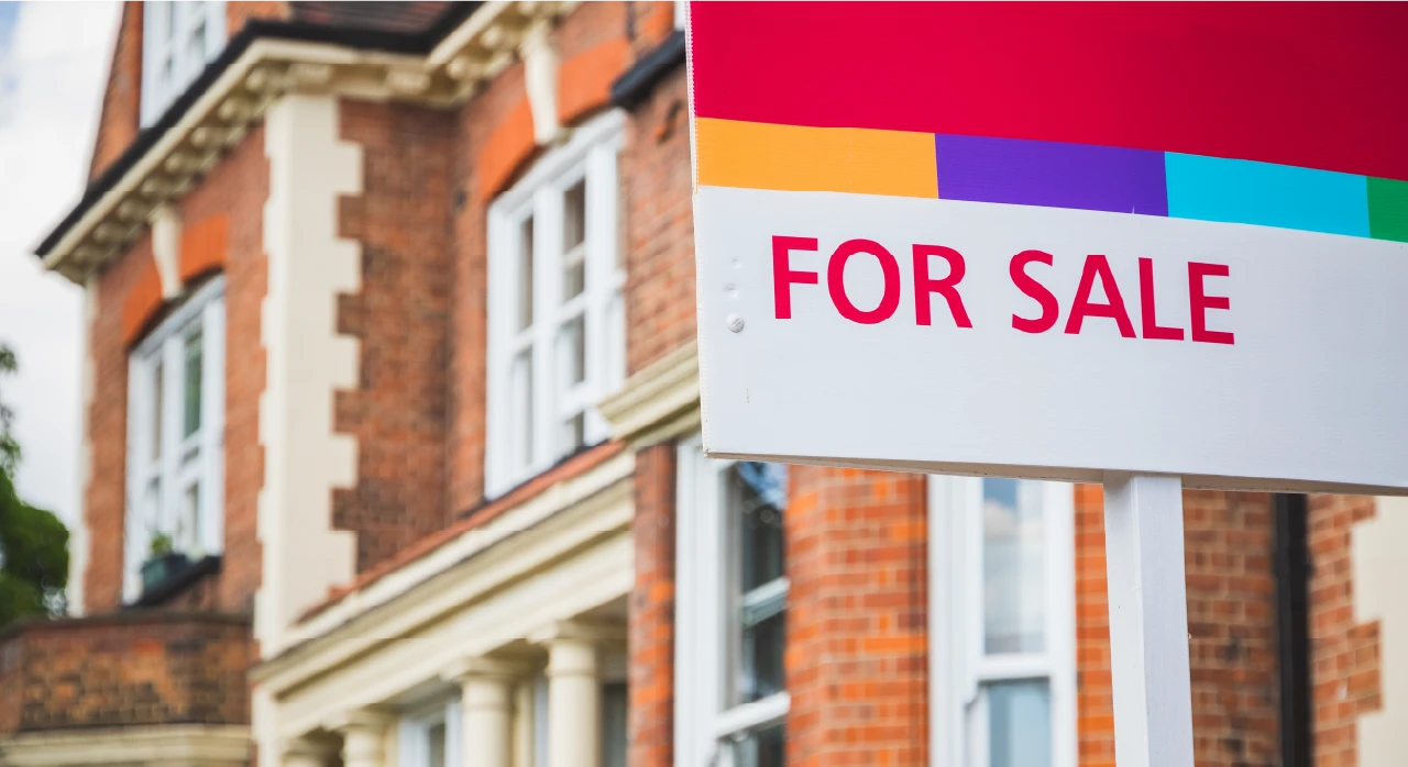 A colourful "For sale" estate agent sign in front of a red-brick period house, representing a UK property sale in progress.